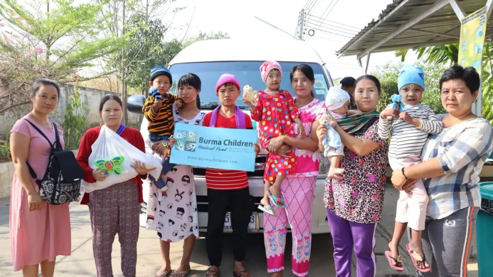 Patients and family members holding a Burma Children Medical Fund banner beside a patient transfer van