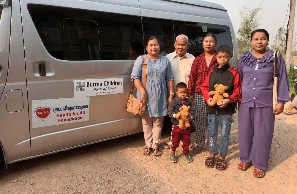 Two young patients and their caregivers stand beside a HFA-BCMF patient van before setting out on the long drive to hospital.