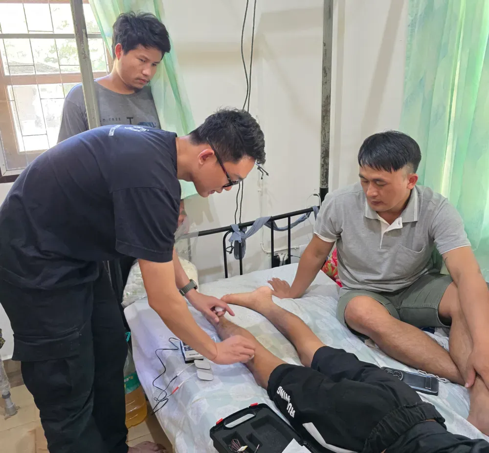 A member of the rehabilitation team places electrodes on a patient's leg during a bedside therapy session, while others look on and learn how the treatment works.